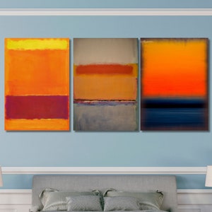 Puede incluir: Tres pinturas abstractas de estilo minimalista, con bandas horizontales de color en tonos de naranja, amarillo, rojo, azul y gris. Las pinturas están colgadas en una pared azul claro sobre una cama tapizada en gris con una manta de lana gris.