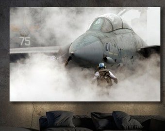 May include: A gray fighter jet taxiing on a runway, with a person in a helmet and flight suit walking in front of it. The jet is surrounded by a cloud of white smoke.