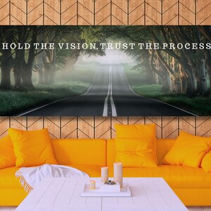 Op de afbeelding: Een rechthoekige canvas print met de tekst "HOLD THE VISION, TRUST THE PROCESS" boven een weg omzoomd door bomen. De print hangt boven een gele bank met bijpassende kussens en een witte salontafel.