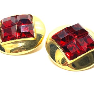May include: A pair of gold-toned earrings with a square, faceted, ruby-red gemstone center. The earrings have a rounded, oval shape with a polished finish. The gemstones are set in a square frame.
