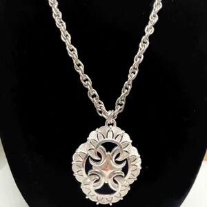 Puede incluir: Collar plateado con un colgante grande. El colgante tiene un diseño floral con una forma central en forma de cruz. La cadena es de diseño grueso tipo cuerda. El collar se muestra sobre un fondo negro.