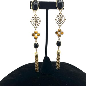 May include: A pair of long, gold-toned earrings with black and amber-colored stones. The earrings feature an oval black stone at the top, a decorative filigree, a cluster of amber stones, another black stone, and a chain tassel. The earrings are displayed on a black stand.
