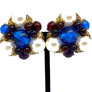 May include: A pair of vintage clip-on earrings. Each earring features a cluster of white faux pearls, blue and red glass beads, and gold-tone metal leaf accents. The earrings are displayed on a black stand against a white background.