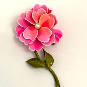 May include: A floral brooch featuring a layered flower design in shades of pink and orange, with a pearl center. The brooch has a green stem and leaves, creating a nature-inspired accessory.