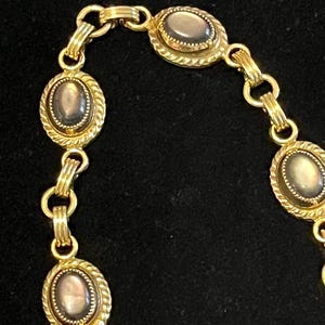 Puede incluir: Un collar dorado con piedras ovaladas e iridiscentes engastadas en marcos texturizados. Los eslabones de la cadena también son dorados, creando un diseño elegante. El collar se muestra sobre un fondo oscuro.