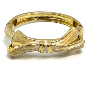 May include: A gold-toned hinged bracelet with a bow design. The bow features a cream-colored enamel finish and is centered on the bracelet. The bracelet has a polished, reflective surface.