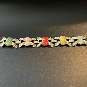 Könnte beinhalten: Ein goldfarbenes Armband mit einer Reihe von farbigen, oval geformten Steinen. Die Steine sind in den Farbtönen Grün, Rosa, Rot, Gelb und Blau gehalten. Das Armband hat einen Verschluss. Das Armband ist ca. 18 cm lang.