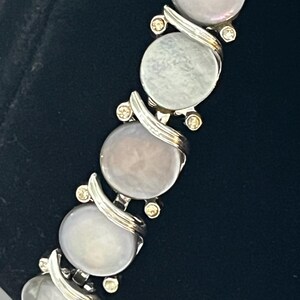 May include: A silver-toned bracelet featuring large, oval, iridescent, mother-of-pearl-like stones. Each stone is connected by a silver swirl design and accented with small, round, clear crystals. The bracelet is displayed against a dark background.