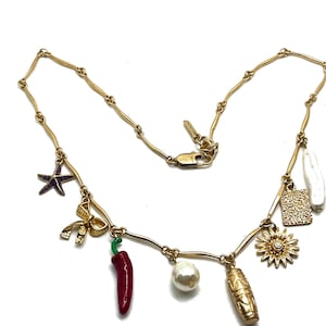 Puede incluir: Collar dorado con varios colgantes, incluyendo un chile rojo, una perla, una estrella de mar, un lazo, un sol, un rectángulo texturizado y un colgante oblongo blanco. El collar tiene un diseño de cadena delicado.