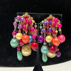 Pode incluir: Um par de brincos de lustre com uma cascata de contas coloridas. Os brincos apresentam tops de filigrana dourada e uma mistura de contas rosa, roxas, laranja, azuis e creme. Os brincos são exibidos em uma superfície de veludo preto.