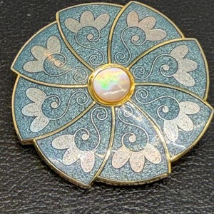May include: A round, blue and gold brooch with a floral design. The brooch features a central opal-like stone and is accented with silver-colored floral patterns. The brooch is set against a black background.