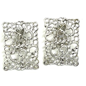 May include: A pair of silver-toned rectangular clip-on earrings. Each earring features an openwork design with swirling patterns and a central teardrop-shaped element. The earrings are set against a white background.