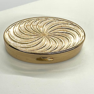 Könnte beinhalten: Goldfarbene ovale Pillendose mit einem Spiralmuster auf dem Deckel. Die Pillendose hat eine aufklappbare Öffnung und eine glatte, metallische Oberfläche. Das Design auf dem Deckel zeigt geschwungene Linien, die von der Mitte ausgehen.
