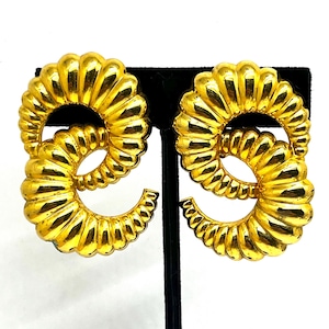 May include: A pair of gold-toned, textured hoop earrings. Each earring features a double-loop design with a ridged, fluted surface. The earrings are displayed on a black stand against a white background.