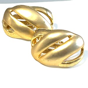 May include: A pair of gold-toned earrings. Each earring has a rounded, cage-like design with curved, solid bars and open spaces. The earrings have a matte finish and are set against a white background.