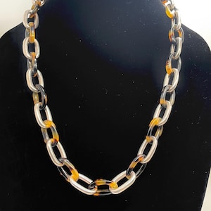 Puede incluir: Un collar de cadena gruesa con eslabones plateados y carey alternados. El collar se exhibe sobre un fondo negro, mostrando su diseño audaz y contraste de color. Un accesorio de moda.