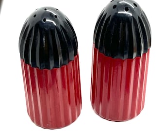 Peut inclure: Paire de salières et poivrières vintage. Les salières et poivrières ont une base rouge côtelée et un dessus noir strié avec plusieurs petits trous. Les salières et poivrières sont de forme cylindrique et reposent sur une surface blanche.