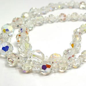 May include: Close-up of a clear crystal beaded necklace. The beads are faceted, reflecting light with iridescent colors. The necklace is on a white background, showcasing the intricate details of the jewelry.
