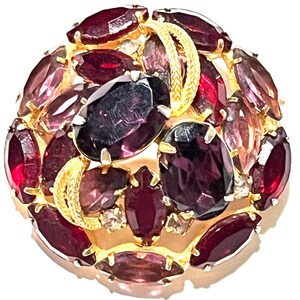 Könnte beinhalten: Eine große, runde Brosche mit einem Cluster aus roten, violetten und klaren Strasssteinen, die in goldfarbenem Metall gefasst sind. Die Strasssteine sind in einem wirbelnden Muster angeordnet.