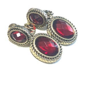 May include: Four pieces of jewelry with oval, faceted, red gemstones. The gemstones are set in a gold-colored metal with a braided rope design around the edges. The jewelry is on a white background.