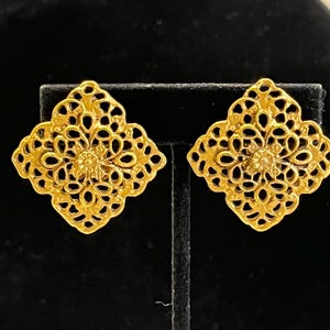 Può includere: Un paio di orecchini in filigrana color oro. Ogni orecchino presenta un design a forma di diamante con un motivo traforato e un centro floreale. Gli orecchini sono esposti su un supporto nero su sfondo nero.