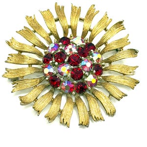 May include: A gold-toned metal flower brooch with a central cluster of red and iridescent rhinestones. The brooch has a sunburst design with elongated, textured petals radiating outwards. The rhinestones are round and set closely together.