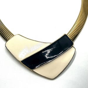 Op de afbeelding: Een vintage goudkleurige ketting met een geometrische hanger. De hanger heeft een zwart en crème emaille ontwerp. De ketting heeft een flexibele, geribbelde ketting. Een stijlvol accessoire.