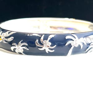 May include: A silver-toned hinged bangle bracelet with a dark blue enamel finish. The bracelet features silver-colored palm tree designs with clear crystal accents. The bracelet is a stylish accessory.