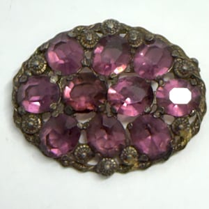 May include: An oval-shaped brooch featuring nine faceted purple stones set in a dark, tarnished metal frame. The metalwork displays a decorative, textured border with small circular accents surrounding the central cluster of oval purple gems.