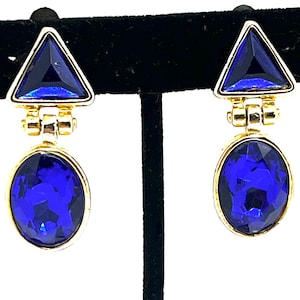 May include: A pair of gold-toned earrings with a geometric design. Each earring features a triangular blue gemstone at the top and an oval blue gemstone at the bottom. The gemstones are faceted, and the earrings are displayed against a black background.