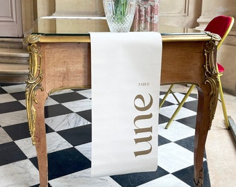 Puede incluir: Una mesa de madera con detalles dorados y una superficie de vidrio. Un camino de mesa blanco con la palabra "une" en letras doradas está sobre ella. Un jarrón transparente con flores blancas está sobre la mesa, con un busto de mármol al fondo.