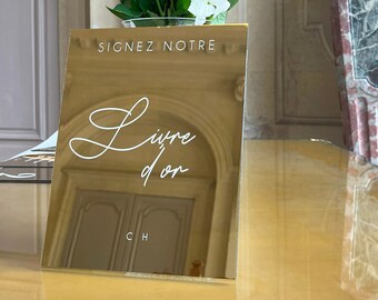 May include: A gold-colored book with the words "Signez Notre Livre d'or" in white script. A bouquet of white flowers sits behind the book. A marble sculpture is in the background.