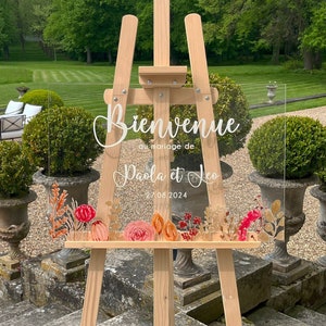 Könnte beinhalten: Ein klares Acryl-Hochzeitschild mit dem Text "Bienvenue au mariage de Paola et Leo 27.08.2024" in weißen Buchstaben. Das Schild ist mit einem floralen Design in Rosa-, Orange- und Goldtönen verziert. Das Schild steht auf einem Holzstaffelei.