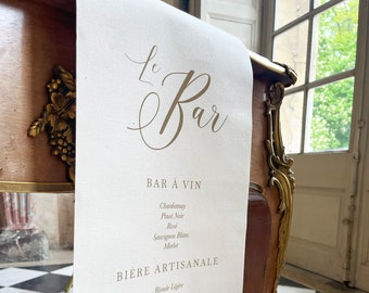 Puede incluir: Un camino de mesa de lino blanco con letras doradas que dicen "Le Bar" y una lista de vinos, cervezas y licores. El menú incluye Chardonnay, Pinot Noir, Rosé, Sauvignon Blanc, Merlot, Blonde Légère, IPA Fratée, Ambrée Equilibrée, Stout Robuste, Blanche Rafraichissante, Vodka Premium, Gin Artisanal, Whisky Single Malt, Ron Vieil y Tequila Reposado.