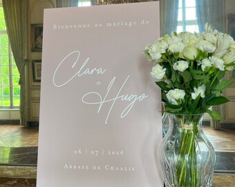 May include: A rectangular dusty pink wedding welcome sign with white text reading Bienvenue au mariage de Clara et Hugo, the date 06 | 07 | 2024, and Abbaye de Chaalis, positioned next to a clear glass vase filled with white roses and carnations.