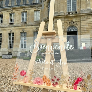 May include: A clear acrylic wedding sign with the text "Bienvenue au mariage de Paola et Leo 27.08.2024" in white calligraphy. The sign is decorated with watercolor floral illustrations in shades of pink, orange, and red, and is displayed on a wooden easel.