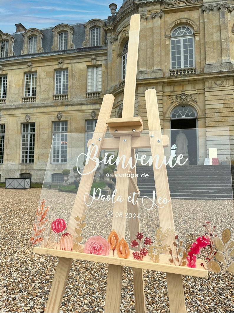 May include: A clear acrylic wedding sign with the text "Bienvenue au mariage de Paola et Leo 27.08.2024" in white calligraphy. The sign is decorated with watercolor floral illustrations in shades of pink, orange, and red, and is displayed on a wooden easel.