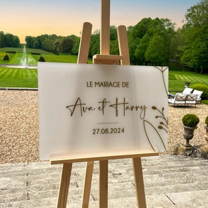 May include: White acrylic wedding sign with gold lettering on an easel. The sign reads "Le Mariage de Ava et Harry 27.08.2024".