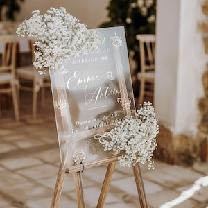 Op de afbeelding: Een transparant acryl huwelijksbord met witte kalligrafie die het huwelijk van Emma en Antoine aankondigt. Het bord staat op een ezel en is versierd met witte baby's breath bloemen. Het bord luidt "Bienvenue au mariage de Emma et Antoine Domaine du Lac Le 10 juillet 2022".