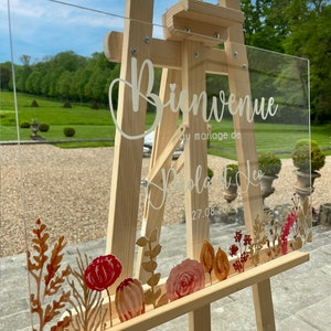 May include: A clear acrylic wedding welcome sign with white calligraphy that reads "Bienvenue au mariage de Pola et Leo 27.08.2024". The sign is decorated with watercolor floral illustrations in shades of red, orange, and brown.