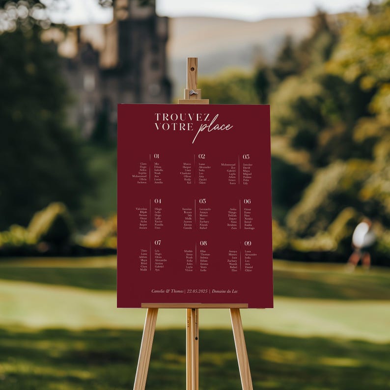 May include: A burgundy wedding seating chart on an easel with the text "TROUVEZ VOTRE place" in white script. The chart lists guest names and table numbers.