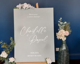 May include: A rectangular sage green wedding welcome sign on a wooden easel. The sign features white text reading Bienvenue à notre mariage, the names Christelle and Pascal in script, the date 29.06.2024, and the location BISTROT DE PARIS.