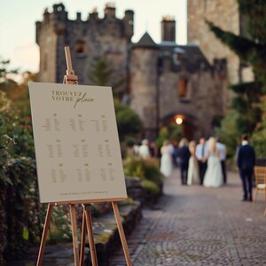 May include: A wedding seating chart on a wooden easel with a beige background and gold text. The chart is titled "Trouvez Votre Place" and lists table numbers and guest names.
