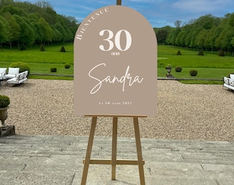 May include: A light brown arch-shaped sign with white text that reads "Bienvenue 30 ans Sandra Le 20 Mars 2027" on a wooden easel.