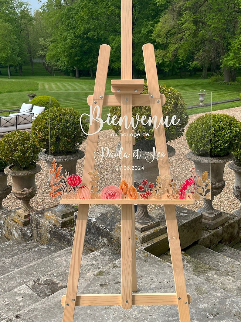 May include: A clear acrylic wedding sign with the text "Bienvenue au mariage de Paola et Leo 27.08.2024" in white lettering. The sign is decorated with a floral design in shades of pink, orange, and gold. The sign is displayed on a wooden easel.