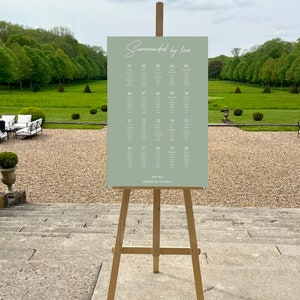 May include: A sage green rectangular wedding seating chart sign on a wooden tripod easel. The sign features the text "Surrounded by love" at the top, followed by a numbered list of twenty tables with guest names, and "20.09.2025 DOMAINE DE CHANTILLY" at the bottom.