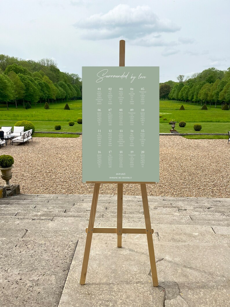 May include: A sage green rectangular wedding seating chart sign on a wooden tripod easel. The sign features the text "Surrounded by love" at the top, followed by a numbered list of twenty tables with guest names, and "20.09.2025 DOMAINE DE CHANTILLY" at the bottom.