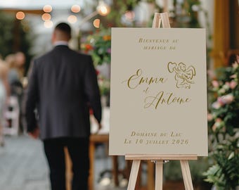 May include: A rectangular beige wedding welcome sign displayed on a light wood easel. The sign features gold text reading Bienvenue au mariage de Emma et Antoine, Domaine du Lac, Le 10 juillet 2026, and a gold line drawing of a cherub holding a heart.