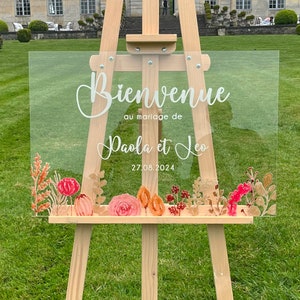 May include: A clear acrylic wedding sign with the text "Bienvenue au mariage de Paola et Leo 27.08.2024" in white lettering. The sign is decorated with watercolor-style flowers in shades of pink, orange, and red.