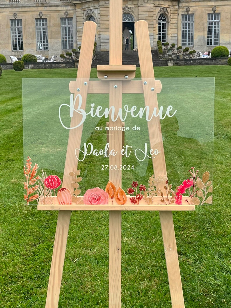 May include: A clear acrylic wedding sign with the text "Bienvenue au mariage de Paola et Leo 27.08.2024" in white lettering. The sign is decorated with watercolor-style flowers in shades of pink, orange, and red.